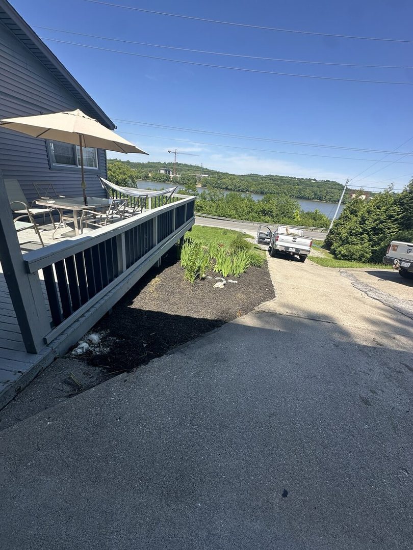 Fresh black mulch bed below a deck, next to the driveway, with a river view behind